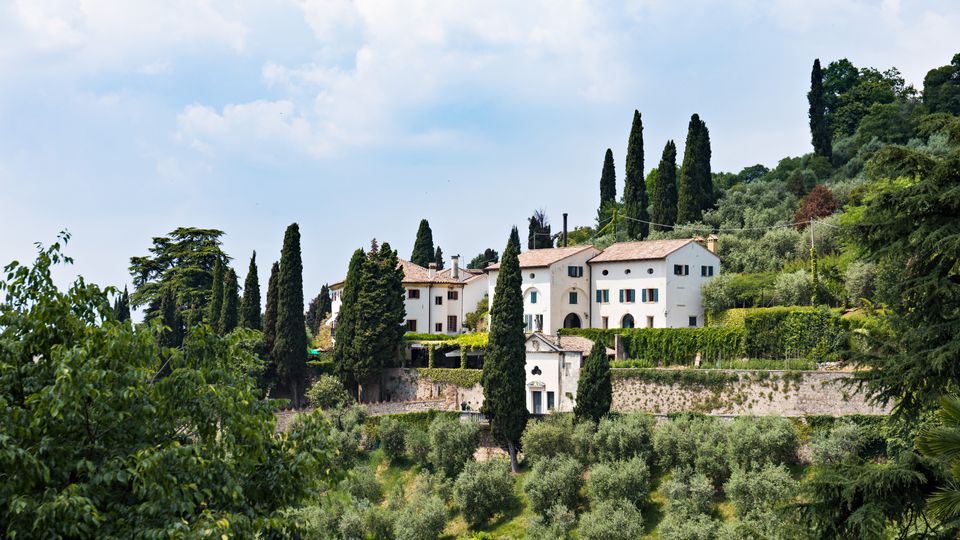 Complesso di edifici rurali in stile villa su una collina, con facciate chiare, tetti in coppi e una piccola cappella. Intorno si vedono cipressi alti, uliveti terrazzati e un muro in pietra, sotto un cielo parzialmente nuvoloso.