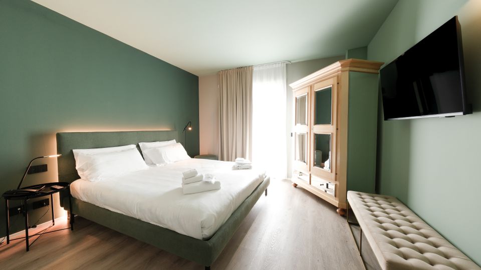 Camera da letto moderna con pareti verde salvia, letto matrimoniale con biancheria bianca e illuminazione a parete. Davanti a una finestra con tende chiare si trovano un armadio in legno con ante a vetro; sulla parete laterale sono presenti una TV a schermo piatto e una panca imbottita.