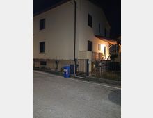 Esterno notturno di una casa a due piani con facciata chiara e finestre scure, circondata da una recinzione metallica con cancello. A destra si vede l’ingresso illuminato con piccolo portico in legno e una scalinata che sale dal marciapiede.