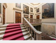 Interno di un palazzo storico con ampia scalinata in pietra rivestita da passatoia rossa e balaustra in marmo che conduce a un pianerottolo. Le pareti chiare sono decorate con grandi dipinti incorniciati e porte in legno scuro, sotto un soffitto alto con cornici modanate.