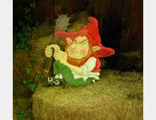 Decorazione dipinta a forma di folletto con cappello rosso appoggiata su balle di fieno, davanti a una parete di assi di legno. Intorno si vede vegetazione rampicante e un’ambientazione rustica da fienile o cortile agricolo.