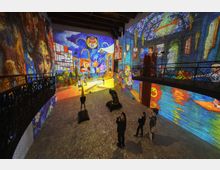 Ampia sala espositiva con soffitto a travi e balconate laterali in ferro battuto, con pavimento in pietra. Pareti interamente coperte da proiezioni artistiche colorate in stile pittorico che trasformano lo spazio in un’installazione immersiva.