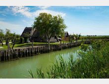 Casa rurale con tetto in paglia affacciata su un canale, con argini rinforzati da pali di legno e vegetazione di canneti in primo piano. Sullo sfondo si vedono prati e alberi in un paesaggio pianeggiante sotto un cielo parzialmente nuvoloso.