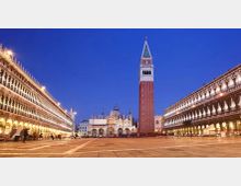 Veduta serale di una grande piazza lastricata con un alto campanile in mattoni al centro e una basilica con cupole sullo sfondo. Ai lati si estendono lunghi edifici porticati illuminati, che incorniciano lo spazio aperto sotto un cielo blu.