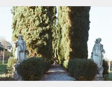 Viale di giardino con sentiero in ghiaia fiancheggiato da alte siepi e cipressi, che crea una prospettiva centrale. Ai lati dell’ingresso del percorso si trovano due statue in pietra su piedistalli tra il verde.