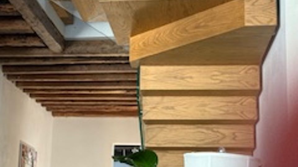 Interno domestico con scala moderna in legno a sbalzo e ringhiera in vetro, sotto un soffitto con travi a vista. Accanto alla scala si vedono una pianta in vaso, un tavolino con lampada e una parete decorata con quadri.