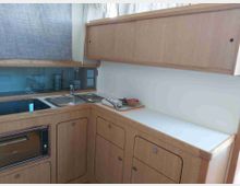 Cucina compatta ad angolo con mobili in legno chiaro e piano di lavoro bianco, dotata di lavello doppio in acciaio e forno integrato. L’area include una piccola finestra e un paraschizzi con pannello grigio e accessori da cucina sul bancone.