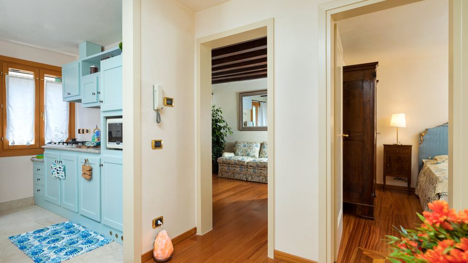 Interno di un appartamento con corridoio in parquet che collega una cucina compatta con mobili azzurri e finestra, un soggiorno con travi a vista e divano, e una camera da letto con armadio scuro e comodino con lampada. Pareti chiare e passaggi tra ambienti senza porte evidenti.