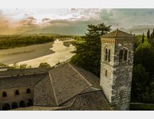 Veduta aerea di un complesso religioso in pietra con tetti in coppi e campanile quadrato con finestre ad arco. Sullo sfondo si vede un ampio fiume che serpeggia nella valle, un ponte in lontananza e montagne sotto un cielo nuvoloso.