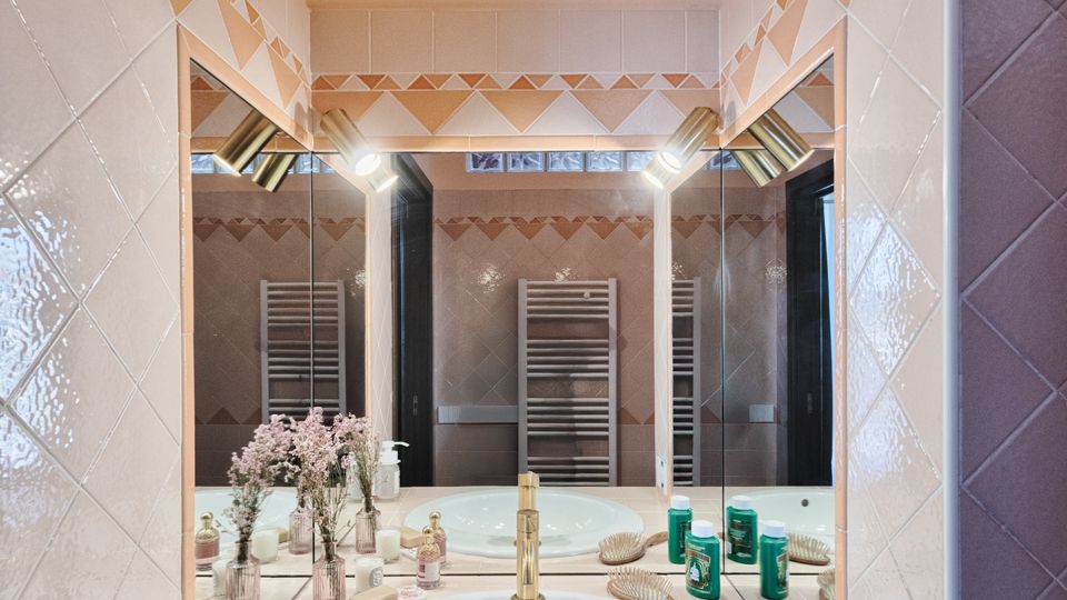 Angolo lavabo di un bagno con ampio specchio incassato, lavandino rotondo e rubinetteria dorata su piano piastrellato chiaro. Pareti rivestite in piastrelle con motivo geometrico e luci laterali, con accessori da toilette e asciugamano sotto il piano.