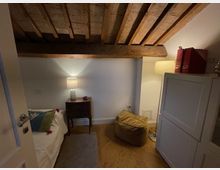 Piccola camera da letto mansardata con soffitto basso e travi in legno a vista, pavimento in legno e pareti chiare. Sono visibili un letto singolo, un comodino con lampada, una lampada da terra e un armadio accanto a un pouf.