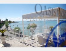 Terrazza panoramica con pergola bianca e ringhiera, arredata con tavolo e sedie, affacciata su una laguna con canali navigabili. Sullo sfondo si vedono la riva alberata, edifici storici con cupola e l’orizzonte della città sull’acqua.