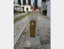 Monumento in pietra con targa in ottone e piccola lampada rossa, collocato lungo un vialetto di ghiaia in un cimitero. Sullo sfondo si vedono cappelle funerarie bianche con croci e alti cipressi, con tombe disposte su prati e camminamenti laterali.