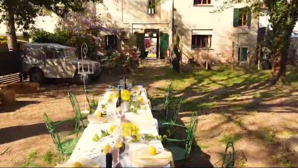 Cortile alberato davanti a un edificio residenziale chiaro, con un lungo tavolo apparecchiato all’aperto e sedie in ferro verde. A sinistra si vede un veicolo d’epoca parcheggiato vicino a una panchina, su terreno misto di ghiaia e prato.