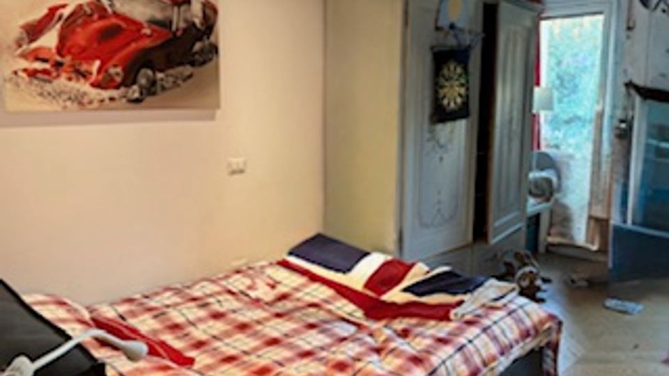 Camera da letto con letto matrimoniale coperto da piumone a quadri e cuscini, pareti chiare con quadro di un’auto rossa. Sullo sfondo ci sono un armadio, una porta e una portafinestra che dà su un’area esterna, con pavimento chiaro e alcuni oggetti sparsi.