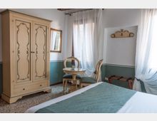 Camera da letto in stile classico con armadio in legno decorato, specchio a parete e piccolo tavolino rotondo con due sedie. Grandi finestre con tende bianche filtrano la luce, mentre un letto con copriletto verde e un portavaligie completano l’arredo.