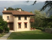 Casa rurale in stile tradizionale con tetto in tegole, pareti color crema e persiane rosse, situata in un ampio giardino con prato verde. Sullo sfondo, si distinguono colline e vegetazione.