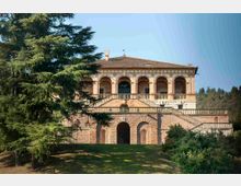 Villa storica su terrazze con facciata ad archi e loggiato superiore, grande scalinata centrale e basamento in pietra con aperture ad arco. Davanti si estende un prato con alberi sempreverdi e vegetazione, sullo sfondo colline boscose sotto un cielo sereno.