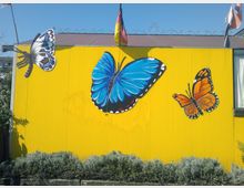 Facciata esterna di un edificio con parete gialla, decorata da grandi murales di farfalle (una blu al centro, una arancione a destra e una bianca a sinistra). In alto si vedono bandiere sul tetto e in basso una fascia di cespugli lungo la base della parete.