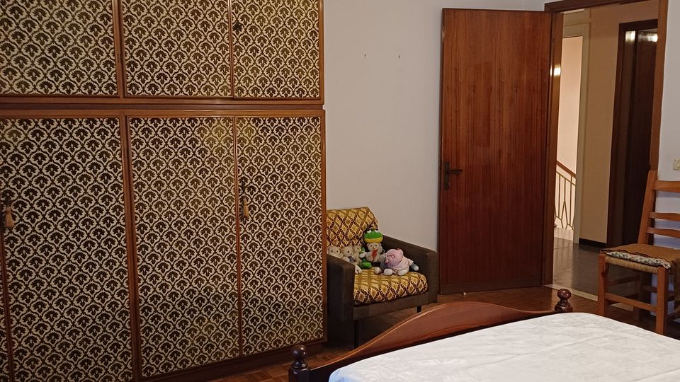 Camera da letto con pavimento in parquet, grande armadio a tutta parete con ante rivestite a motivo geometrico e letto in legno con copriletto bianco. A destra si vedono una porta aperta verso il corridoio, una sedia in legno e un lampadario a soffitto con più luci.