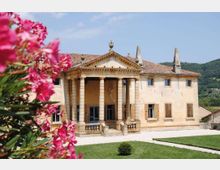 Villa storica in pietra con portico centrale a colonne e frontone triangolare, tetto in coppi e finestre con scuri. Davanti si vede un cortile chiaro e un prato curato, con colline boscose sullo sfondo e fiori rosa in primo piano.