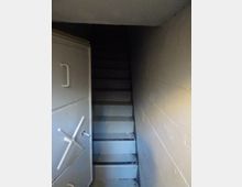 Scala stretta in cemento grigio che scende in un vano sotterraneo dalle pareti lisce, con illuminazione fioca. A sinistra si vede una pesante porta metallica tipo caveau con maniglia e ruota di chiusura.