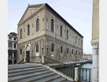 Grande edificio storico in mattoni affacciato su un canale, con facciate scandite da alte finestre ad arco e cornici in laterizio. In primo piano si vedono una scalinata in pietra e una passerella con ringhiera metallica lungo l’acqua.