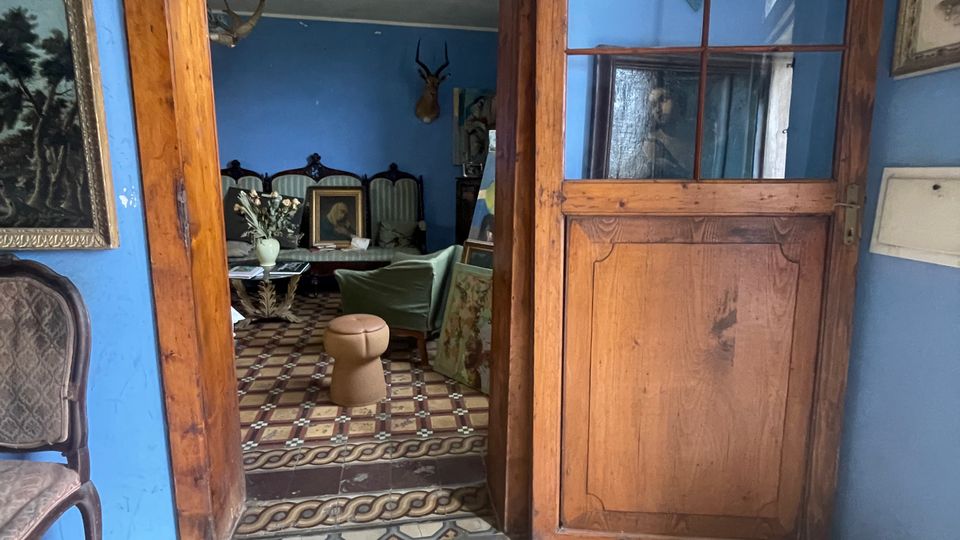 Interno di una casa d’epoca con pareti azzurre, pavimento a piastrelle geometriche e porta in legno con vetri che conduce a un salotto arredato. Sulle pareti sono appesi quadri e una testa imbalsamata, e nella stanza oltre l’ingresso si vedono mobili e decorazioni in stile vintage.