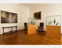 Sala museale con pareti bianche e pavimento in parquet a spina di pesce, con due vetrine su piedistalli in legno che espongono piccole sculture. Alle pareti sono appesi grandi dipinti incorniciati e lungo i lati si vedono un tavolo consolle e alcune sedie d’epoca.