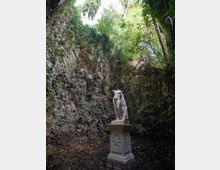Radura in una piccola gola rocciosa ricoperta di edera e vegetazione, con pareti di pietra ripide e alberi che sovrastano l’apertura. Al centro si trova un piedistallo in pietra decorato con motivi a stella, sormontato da una statua classica in marmo.