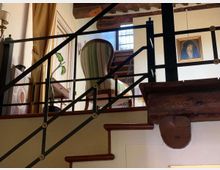 Interno di una casa con scala in legno e ringhiera in ferro che porta a un soppalco arredato con poltrona a righe verdi, tavolino e pianta. Soffitto con travi a vista, finestra con vetri decorati, tende e un quadro appeso alla parete.