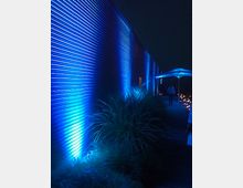 Corridoio esterno notturno lungo un edificio con parete in metallo ondulato illuminata da luci blu dal basso. Vegetazione ornamentale sul lato e, in fondo, una piccola struttura coperta con ulteriori punti luce.