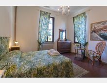 Camera da letto luminosa con pareti bianche, due finestre alte con tende a motivo tropicale e letto matrimoniale coordinato con biancheria verde. Arredo con comò in legno e specchio, lampadario a soffitto, tavolino e sedia in vimini, tappeto e quadro a parete.