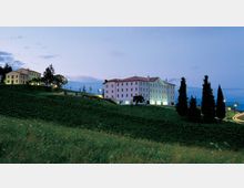 Grande edificio in stile rurale con facciata chiara e tetto in tegole rosse su una collina erbosa, con finestre illuminate al crepuscolo. Intorno si vedono campi e alberi slanciati, alcune luci lungo i vialetti e una strada curva sul lato destro.