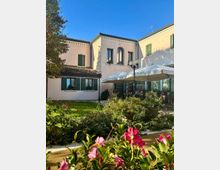 Cortile-giardino di un edificio a due piani intonacato chiaro, con finestre ad arco e persiane verdi e un tetto in coppi. In primo piano aiuole con fiori rosa e, sulla destra, un’area coperta da grandi ombrelloni bianchi con lampioni e tavoli all’esterno.