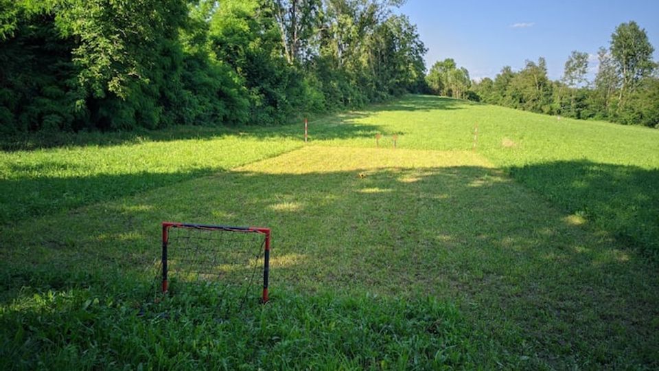 Ampio prato erboso in una radura, circondato da alberi e vegetazione fitta ai lati, con cielo sereno sullo sfondo. In primo piano si vede una piccola porta da gioco e, più lontano, alcuni paletti che delimitano un’area sul terreno.