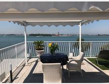 Terrazza panoramica coperta da tenda bianca con ringhiera in metallo, tavolo con tovaglia scura e sedie in rattan. Sullo sfondo si vede una laguna con barche e un’isola con edifici e campanili all’orizzonte.