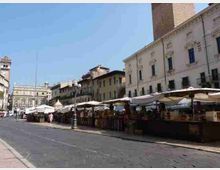 Piazza cittadina con fila di bancarelle di mercato sotto grandi ombrelloni lungo il bordo della strada. Sullo sfondo si vedono edifici storici con facciate chiare e una torre, con pavimentazione in pietra e cielo sereno.