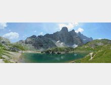 Lago alpino dalle acque turchesi in una conca montana, circondato da pendii erbosi e rocce grigie. Sullo sfondo si innalzano alte cime rocciose frastagliate con piccoli residui di neve sotto un cielo azzurro con nuvole sparse.
