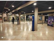 Atrio interno di un cinema o centro eventi con pavimento in pietra chiara, colonne cilindriche e controsoffitto scuro a griglia con luci incassate. Sullo sfondo si vedono biglietterie, vetrate con decorazioni e poster cinematografici con transenne per le file.