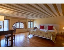 Camera da letto in stile tradizionale con pavimento in legno, soffitto a travi dipinte e ampie finestre ad arco con vetri decorati. Al centro c’è un letto matrimoniale con copriletto floreale e cuscini rossi, affiancato da comodini con lampade, e un tavolo con sedie vicino alla finestra.