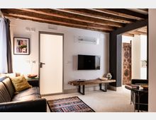 Soggiorno di un appartamento con soffitto a travi di legno a vista, pareti bianche e pavimento chiaro. Divano in pelle scura, tappeto colorato, TV a parete e panca in legno rustico, con apertura verso una zona cucina e pannello decorativo intagliato.