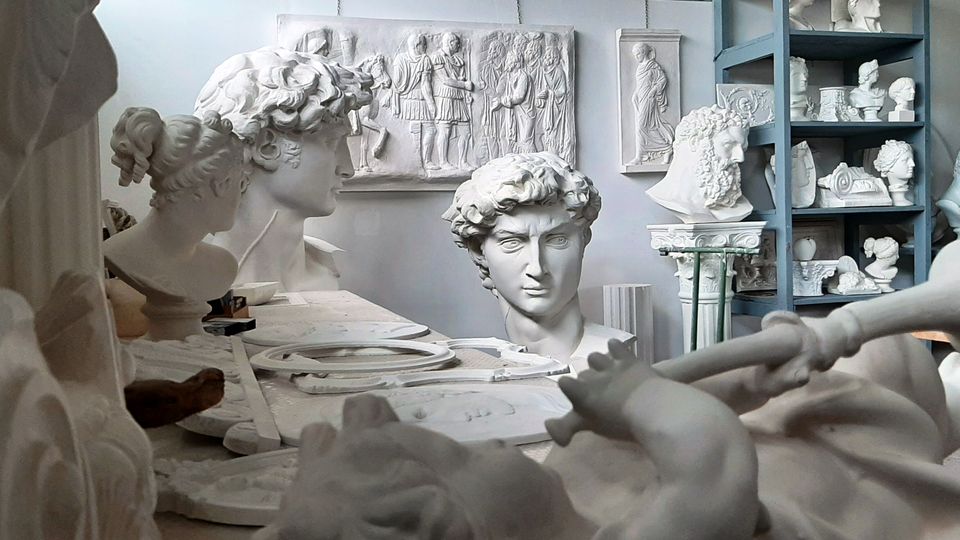 Interno di un laboratorio di scultura pieno di calchi in gesso, con busti classici su un tavolo e su scaffalature. Sullo sfondo si vedono rilievi e statue appoggiate alle pareti in un ambiente luminoso e affollato di opere.