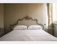 Camera da letto con letto matrimoniale dai tessili bianchi e testata decorativa in stile barocco in metallo. Ai lati ci sono due piccoli tavolini rotondi, e a destra una finestra con tende chiare che illumina la stanza.
