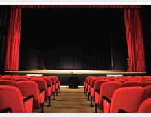 Interno di un teatro con file di poltrone rosse e una corsia centrale che conduce al palcoscenico. Il palco è incorniciato da tende rosse laterali e superiori, con fondale scuro e sipario nero.