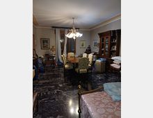 Ampia sala da pranzo in stile tradizionale con pavimento in marmo scuro, grande tavolo in legno con sedie imbottite e lampadario centrale. Alle pareti ci sono finestre con tende, quadri, un’alta credenza vetrata e vari mobili e oggetti domestici.