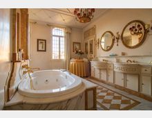 Ampio bagno in stile classico con vasca idromassaggio incassata in marmo e rubinetteria dorata. Lungo la parete ci sono doppi lavabi con mobili intagliati, specchi ovali, applique e un grande finestrone ad arco che illumina il pavimento a scacchi.