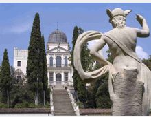 Statua in pietra in primo piano che domina un lungo viale di scalinate, fiancheggiato da alti cipressi, che conduce a una villa neoclassica bianca con facciata colonnata e cupola. Sullo sfondo si vedono giardini alberati e cielo sereno.