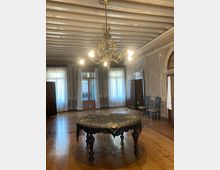 Salone storico con pavimento in legno e soffitto a travi dipinte, illuminato da un grande lampadario in ottone. Al centro c’è un tavolo rotondo con tovaglia ricamata e frange, circondato da pareti ornate e alte finestre con tende bianche.