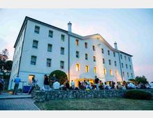 Grande edificio bianco a più piani con facciata semplice, numerose finestre rettangolari e frontone centrale, illuminato da luci calde al crepuscolo. Davanti si trova un terrazzo con muretto in pietra e prato, in un’area esterna tipo cortile o giardino.